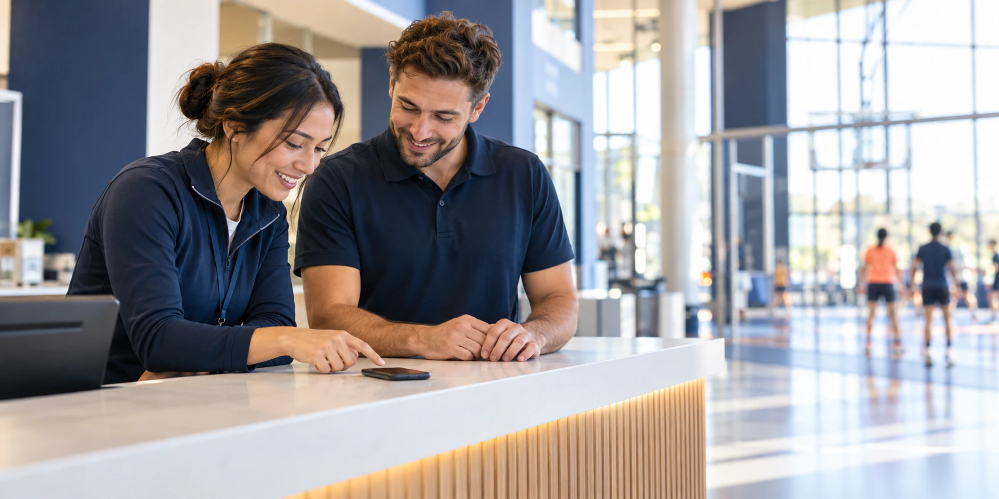 Two service colleagues review an unreadable mobile conversation together at a bright sports-centre reception